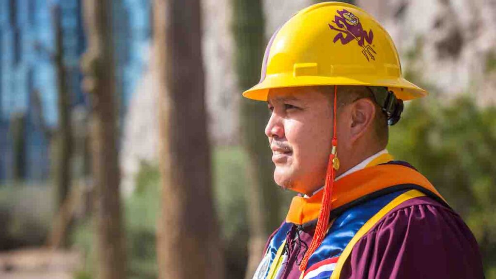 A man in a hard hat and graduation robe stands proudly.