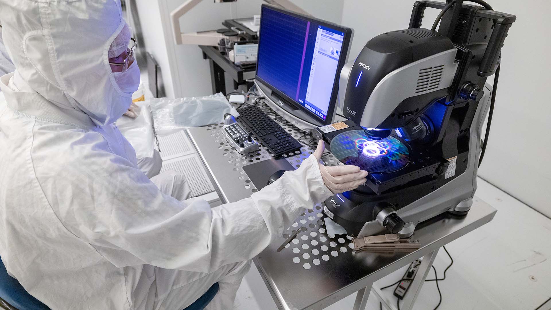 A researcher in a cleanroom suit operates a high-powered microscope to inspect a silicon wafer in a semiconductor lab. The wafer displays iridescent patterns under the microscope, which is connected to a dual-monitor computer system showing inspection software.
