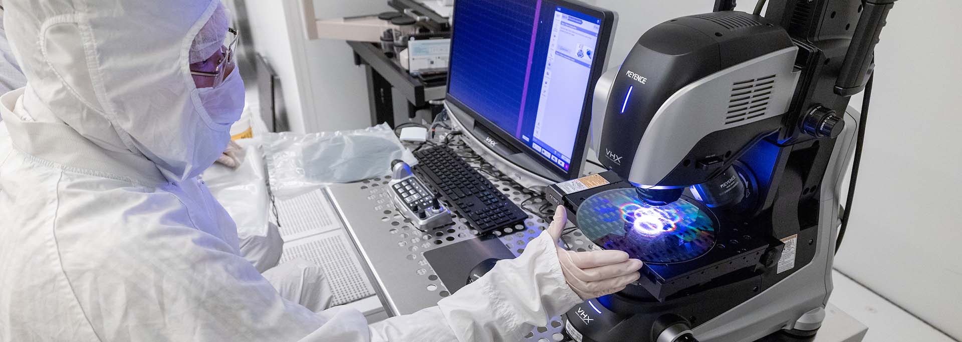 A researcher in a cleanroom suit operates a high-powered microscope to inspect a silicon wafer in a semiconductor lab. The wafer displays iridescent patterns under the microscope, which is connected to a dual-monitor computer system showing inspection software.
