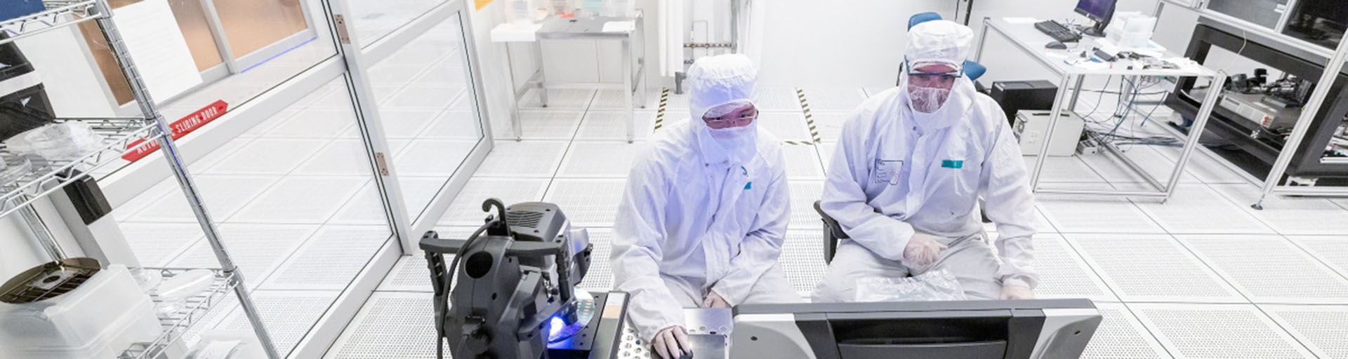 Two individuals in white suits collaborate on a computer, focused on their work in a lab setting.