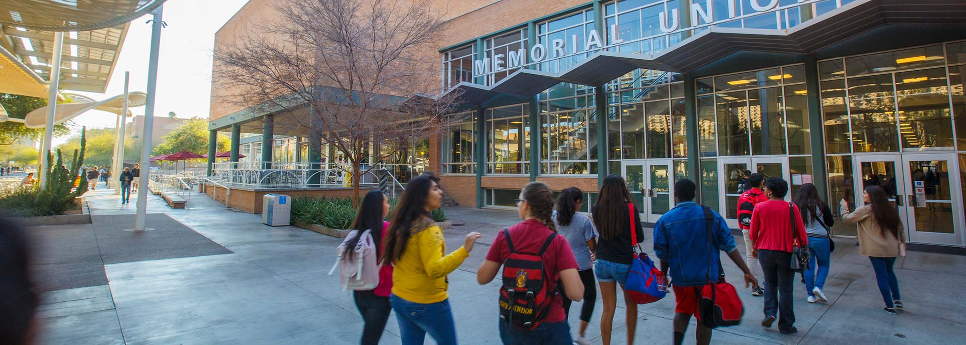 Students carrying backpacks and bags walk toward the entrance of Memorial Union on a university campus.
